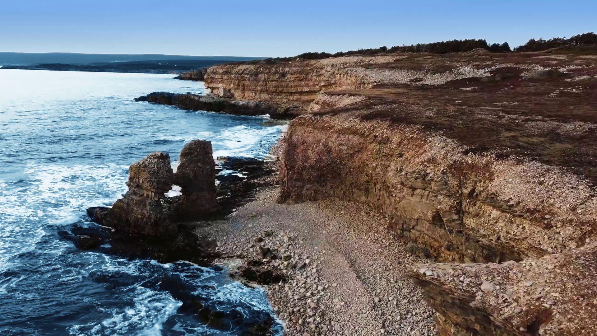Waves breaking below layered cliffs, with a sea stack and a stony beach under a blue sky
