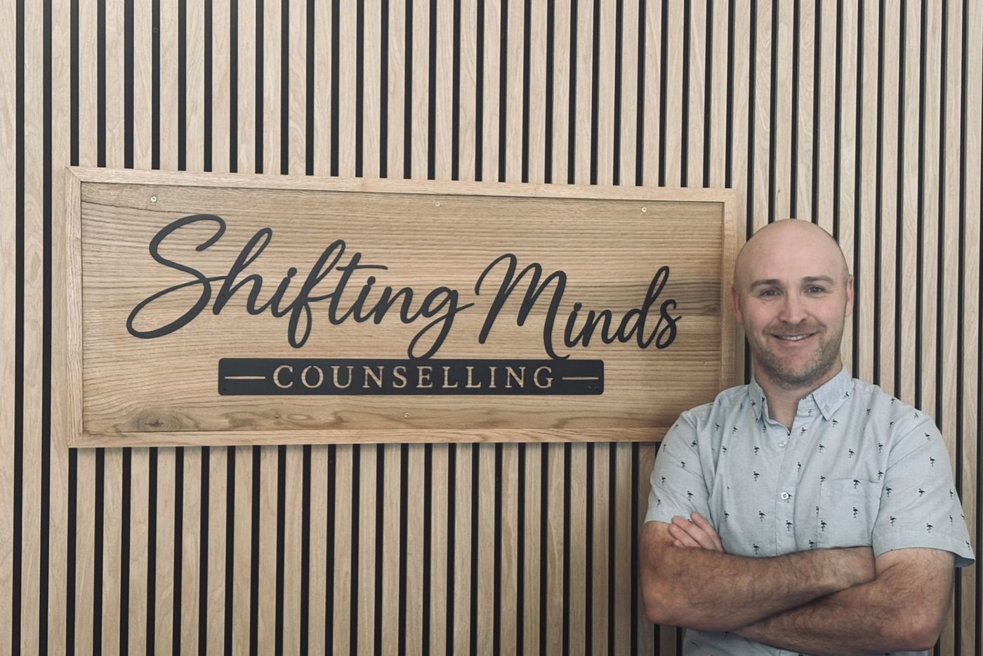 Jimmy Rose, Registered Counselling Therapist, standing with his arms crossed beside the wooden Shifting Minds Counselling sign on his office wall